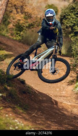 homme méconnaissable dans un casque, des gants et des lunettes de protection sautant faire un tour de fouet en descente pendant la pratique du vtt dans la forêt de bois Banque D'Images