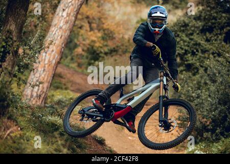 homme méconnaissable dans un casque, des gants et des lunettes de protection sautant faire un tour de fouet en descente pendant la pratique du vtt dans la forêt de bois Banque D'Images