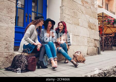 Trois copines utilisant un smartphone dans la rue de la ville. Des femmes élégantes assis près d'un café et s'amuser à bavarder à l'extérieur Banque D'Images