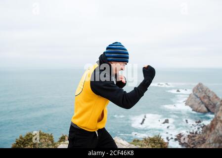 Homme barbu adulte dans les vêtements de sport pratiquant des coups de poing pendant l'entraînement de kickboxing sur une falaise rocheuse près de la mer Banque D'Images