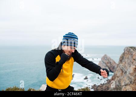 Homme barbu adulte dans les vêtements de sport pratiquant des coups de poing pendant l'entraînement de kickboxing sur une falaise rocheuse près de la mer Banque D'Images