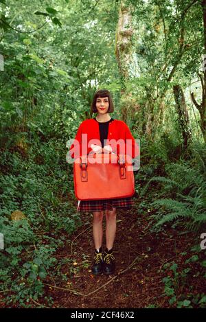Jeune femme élégante en chandail rouge avec grande valise rouge debout regardant directement l'appareil photo dans une forêt dense Banque D'Images
