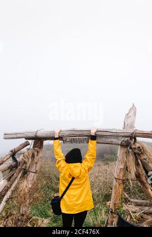 Vue arrière d'une femme perdue non reconnaissable touriste dans un imperméable jaune vif et une capuche avec sac à main pour explorer l'environnement tout en s'appuyant sur une poutre de clôture en bois dans un champ de brouillard avec une mauvaise visibilité Banque D'Images