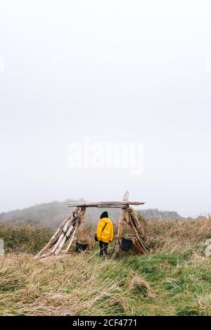 Vue arrière d'une femme perdue non reconnaissable touriste dans un imperméable jaune vif et une capuche avec sac à main pour explorer l'environnement tout en s'appuyant sur une poutre de clôture en bois dans un champ de brouillard avec une mauvaise visibilité Banque D'Images