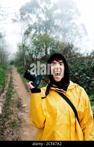 Femme touriste en imperméable jaune vif et capot noir avec un appareil photo numérique criant tout en regardant seul l'appareil photo route parmi la forêt brumeuse Banque D'Images