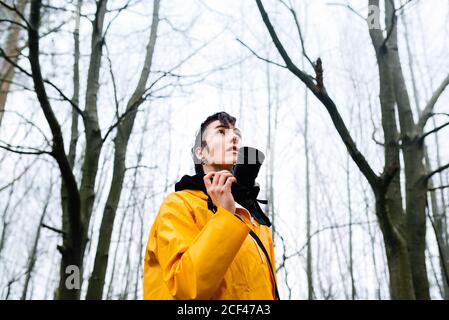 D'en-dessous touristique féminine en imperméable jaune vif et noir cagoule avec appareil photo numérique qui ne se tient pas debout sur le brouillard forêt Banque D'Images