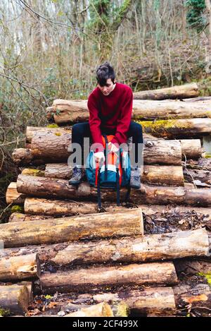 Coupe basse de femme dans des vêtements décontractés et des baskets ouvrant le sac à dos tout en étant assis sur des rondins en bois et en se reposant sur une pente de colline parmi la verdure dans la forêt en journée en Espagne Banque D'Images