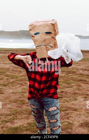 Personne castingmante méconnaissable avec sac en papier et grimace triste sur la tête tenant le sac en plastique et montrant l'affiche de désapprobation pendant debout sur la rive Banque D'Images