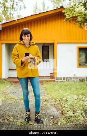 Jeune femme dans une tenue décontractée souriant et parcourant un smartphone pendant debout sur un chemin carrelé à l'extérieur d'un charmant cottage le jour de l'automne en campagne Banque D'Images