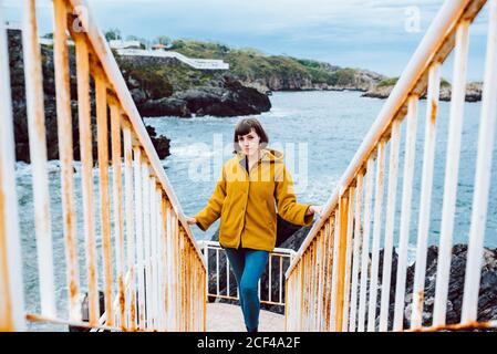 Jeune femme en blouson jaune debout sur un escalier rouillé contre baie avec eau de mer Banque D'Images