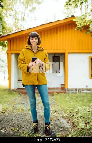 Jeune femme dans une tenue décontractée souriant et parcourant un smartphone pendant debout sur un chemin carrelé à l'extérieur d'un charmant cottage le jour de l'automne en campagne Banque D'Images