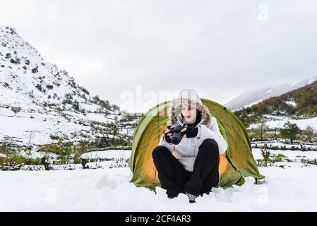 Contenu Femme en blouson d'hiver blanc et pantalon noir assis avec caméra près d'une tente touristique vert clair sur la neige montagnes Banque D'Images