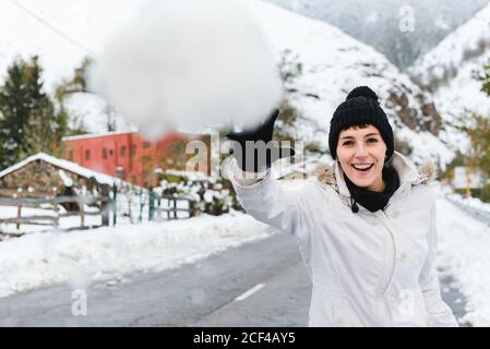 Joyeuse femme en bonnet tricoté noir avec pompon et blanc veste d'hiver qui jette un ballon de neige à la caméra tout en marchant en montagne village Banque D'Images