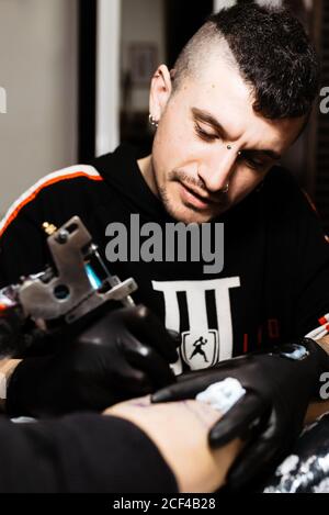 Homme élégant avec perçage en utilisant la machine de tatouage pour faire le tatouage sur la jambe de récolte client pendant le travail dans le salon Banque D'Images