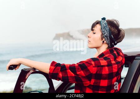 Vue latérale d'une femme extraordinaire avec cheveux courts noirs et bandana sur la tête et piercing dans la chemise à carreaux rouges penchée sur la voiture et regardant loin de la mer Banque D'Images