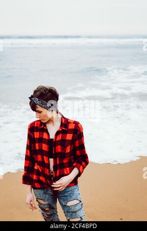 De dessus de la femme pensive malheureuse dans la chemise à carreaux et jean déchiré debout avec une cigarette à la main sur une plage de sable avec la mer de tempête en arrière-plan Banque D'Images