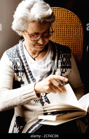 Content femme avec les cheveux gris et sourire gentil lecture livre par fenêtre dans le jour de lumière Banque D'Images