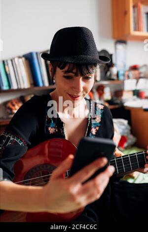 Femme souriante dans un vêtement tendance et chapeau assis sur le lit avec guitare acoustique et lecture de messages sur le téléphone portable tout en refroidissant pendant le week-end Banque D'Images