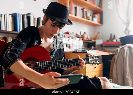 Femme en tenue tendance et chapeau assis sur le lit avec guitare acoustique et lecture de messages sur le téléphone portable tout en refroidissant pendant week-end Banque D'Images