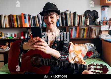 Femme souriante dans un vêtement tendance et chapeau assis sur le lit avec guitare acoustique et lecture de messages sur le téléphone portable tout en refroidissant pendant le week-end Banque D'Images