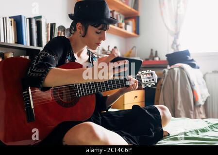 Femme en tenue tendance et chapeau assis sur le lit avec guitare acoustique et téléphone portable tout en se refroidissant pendant le week-end Banque D'Images