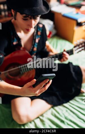 Femme en tenue tendance et chapeau assis sur le lit avec guitare acoustique et lecture de messages sur le téléphone portable tout en refroidissant pendant week-end Banque D'Images