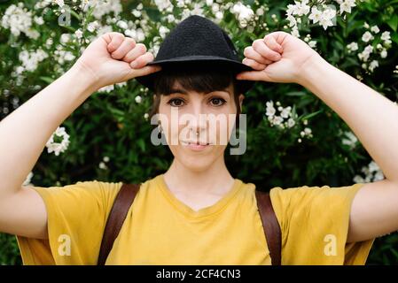 Positive jeune femme élégante dans une chemise jaune décontractée et élégante chapeau noir se tenant près des buissons en fleurs et regardant l'appareil photo Banque D'Images