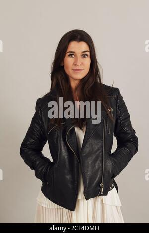 Femme élégante et bien pensée aux cheveux foncés dans une veste de motard noire et regardant l'appareil photo sur fond gris en studio Banque D'Images