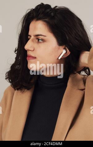 Brunette féminine jeune et confiante en pelage beige avec écouteurs à l'écoute pour écouter de la musique sur fond gris en studio Banque D'Images