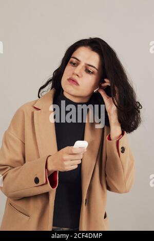 Brunette féminine jeune et confiante en couche beige placée sur les écouteurs et écouter de la musique sur fond gris en studio Banque D'Images