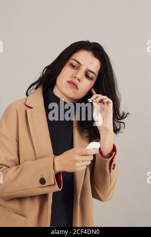 Brunette féminine jeune et confiante en couche beige placée sur les écouteurs et écouter de la musique sur fond gris en studio Banque D'Images