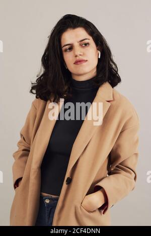 Brunette féminine jeune et confiante en pelage beige avec écouteurs à l'écoute pour écouter de la musique sur fond gris en studio Banque D'Images