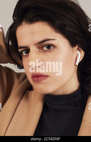 Brunette féminine jeune et confiante en pelage beige avec écouteurs à l'écoute pour écouter de la musique sur fond gris en studio Banque D'Images