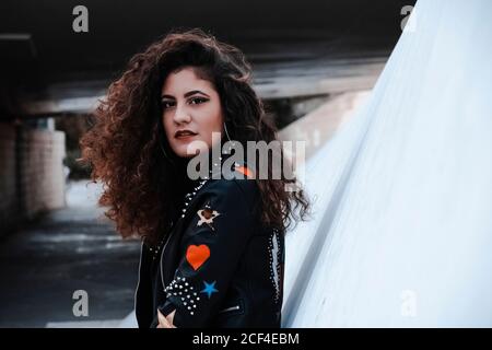 Brunette moderne et confiante avec de longs cheveux ondulés portant du cuir noir veste avec rayures colorées et métalliques touchant les cheveux et le look à la caméra tout en se tenant à côté de la construction en pierre dans la ville Banque D'Images