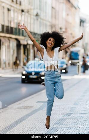 Bonne femme ethnique dans une tenue tendance sautant pour la joie et regardant l'appareil photo le jour de l'été Banque D'Images