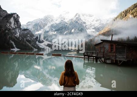 Vue arrière de la femme anonyme voyageur en tenue décontractée debout et se délarant dans la vue de la maison en bois sur le lac turquoise avec les montagnes moggy Dolomites en arrière-plan à l'Italie Banque D'Images