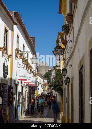 Touristes marchant sur une rue pavée étroite flanquée de petits magasins/magasins dans la ville fortifiée d'Obidos, Estrémadure, Portugal. Banque D'Images