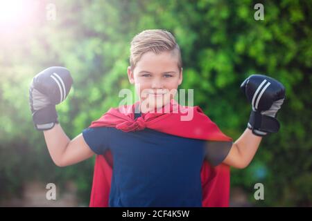 Super-héros de garçon avec des gants de boxe et un manteau rouge montrant de la puissance en extérieur. Concept d'enfance, de réussite et de liberté Banque D'Images