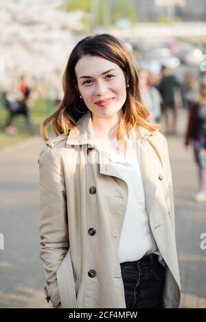 Jolie jeune femme dans un manteau élégant et tendance souriant et regardant l'appareil photo tout en se tenant sur un fond flou de la rue de la ville le jour ensoleillé Banque D'Images