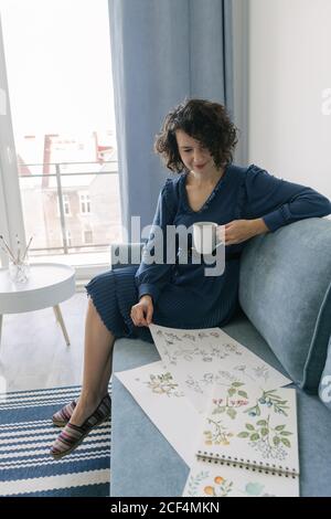 Belle jeune femme brunette assis sur un canapé et un album avec des dessins à la maison tout en dégustant une tasse de café Banque D'Images