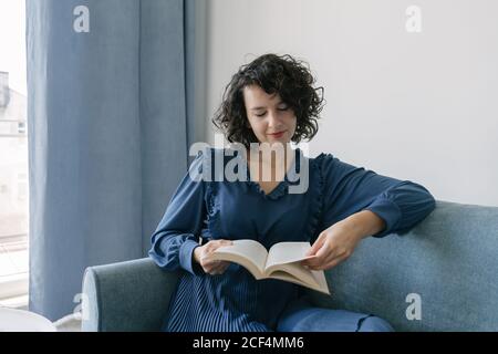 Bonne jeune femme brune en robe bleue livre de lecture assis sur un canapé à la maison Banque D'Images