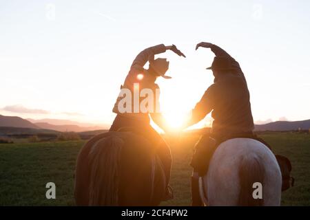 Vue arrière de l'homme et de la femme à cheval et faisant la forme du coeur avec les mains contre le ciel de coucher de soleil sur le ranch Banque D'Images