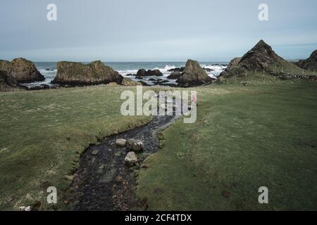 D'en haut, une femme anonyme appréciant le paysage magnifique de l'Irlande du Nord pendant le voyage tout en marchant près de la rivière rapide peu profonde qui coule aux eaux de la rive rocheuse Banque D'Images