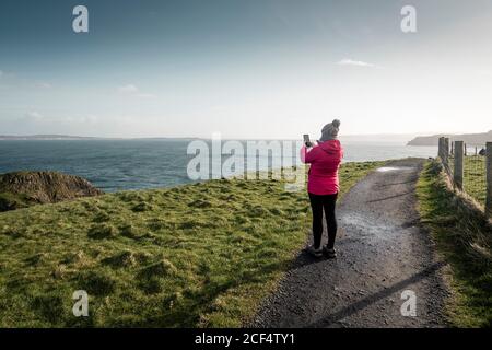 Vue arrière de la femme voyageur dans une chaude activewear debout route rurale longeant le rivage avec de l'herbe verte et de prendre photo de l'océan avec de l'eau calme le jour ensoleillé du printemps En Irlande du Nord Banque D'Images