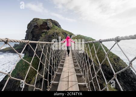 Vue arrière de la touriste femelle en veste rouge qui marche Corde Carric un pont de rede suspendu entre le continent et petit rocky Island sur la côte d'Irlande du Nord Banque D'Images