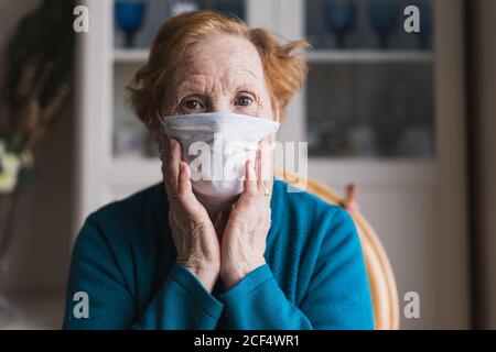 Femme sénior avec cheveux rouges en robe bleue et médical masque regardant la caméra en se tenant dans la chambre d'hôpital Banque D'Images