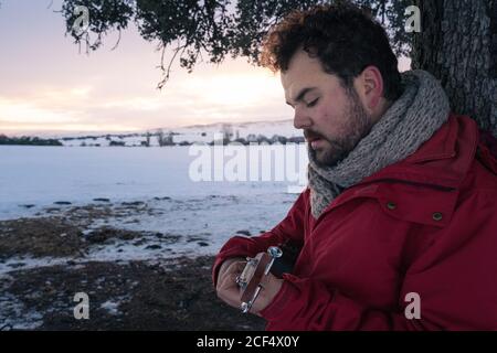 Vue latérale d'un homme d'âge moyen dans des vêtements chauds jouant de la guitare sous l'arbre sur fond de paysage enneigé Banque D'Images