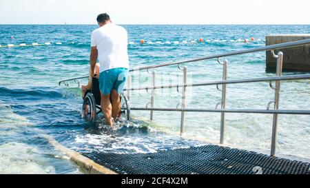 Homme handicapé à la plage nageant sur un fauteuil roulant avec aide sur une rampe d'accès. Banque D'Images