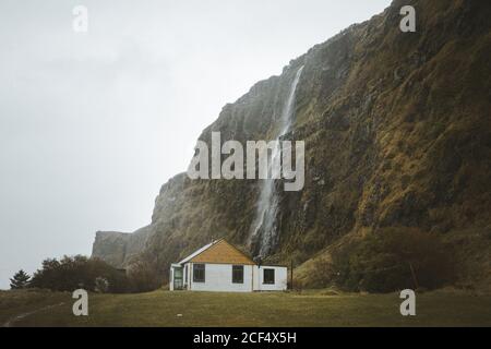 Petite maison de campagne en bois avec murs blancs et pignons jaunes toit situé sur la prairie verte au pied de la falaise avec Cascade contre ciel gris nuageux le jour du printemps dans le Nord Irlande Banque D'Images