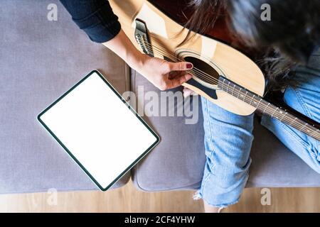 Femme jouant de la guitare assis sur son canapé à la maison et apprendre avec des cours en ligne avec une tablette numérique avec un tablette numérique avec un écran vide ci-dessus Banque D'Images